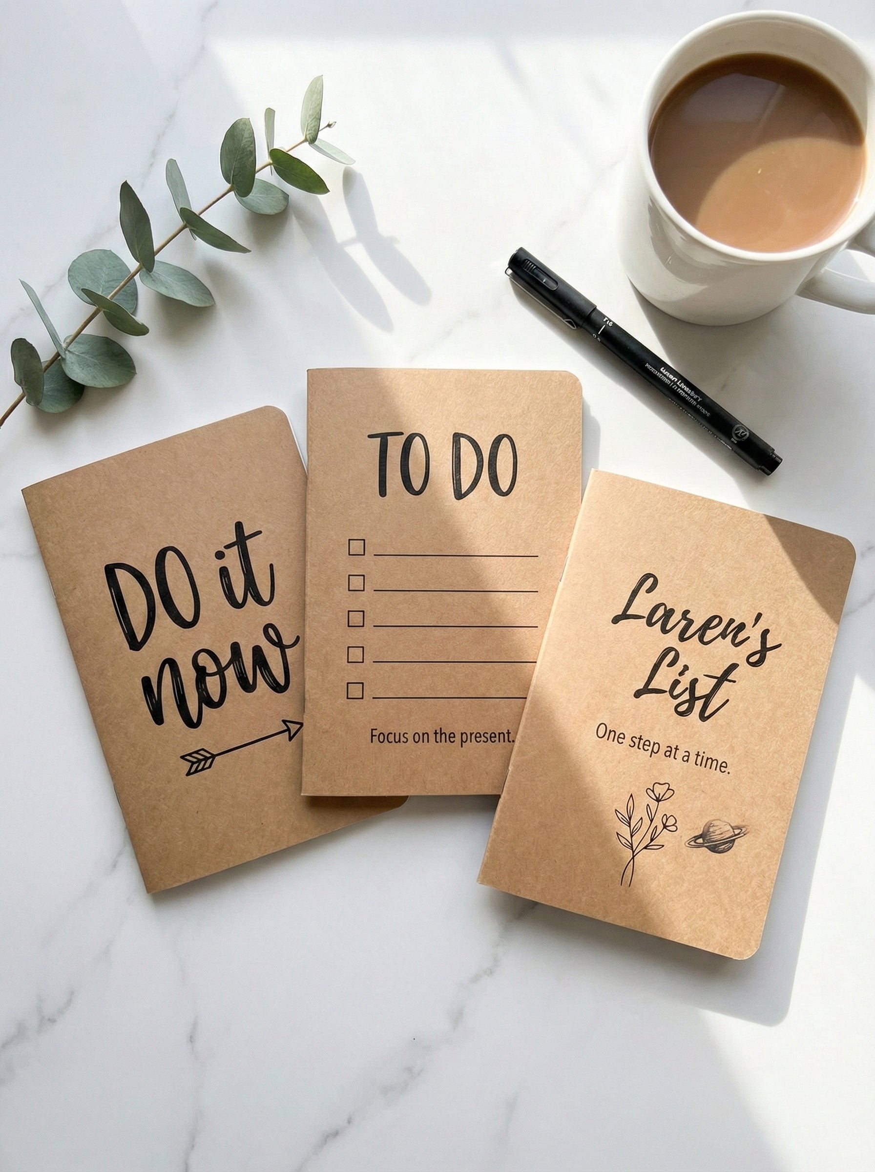 Three small notebooks with motivational phrases on a marble surface with a cup of coffee and eucalyptus leaves.
