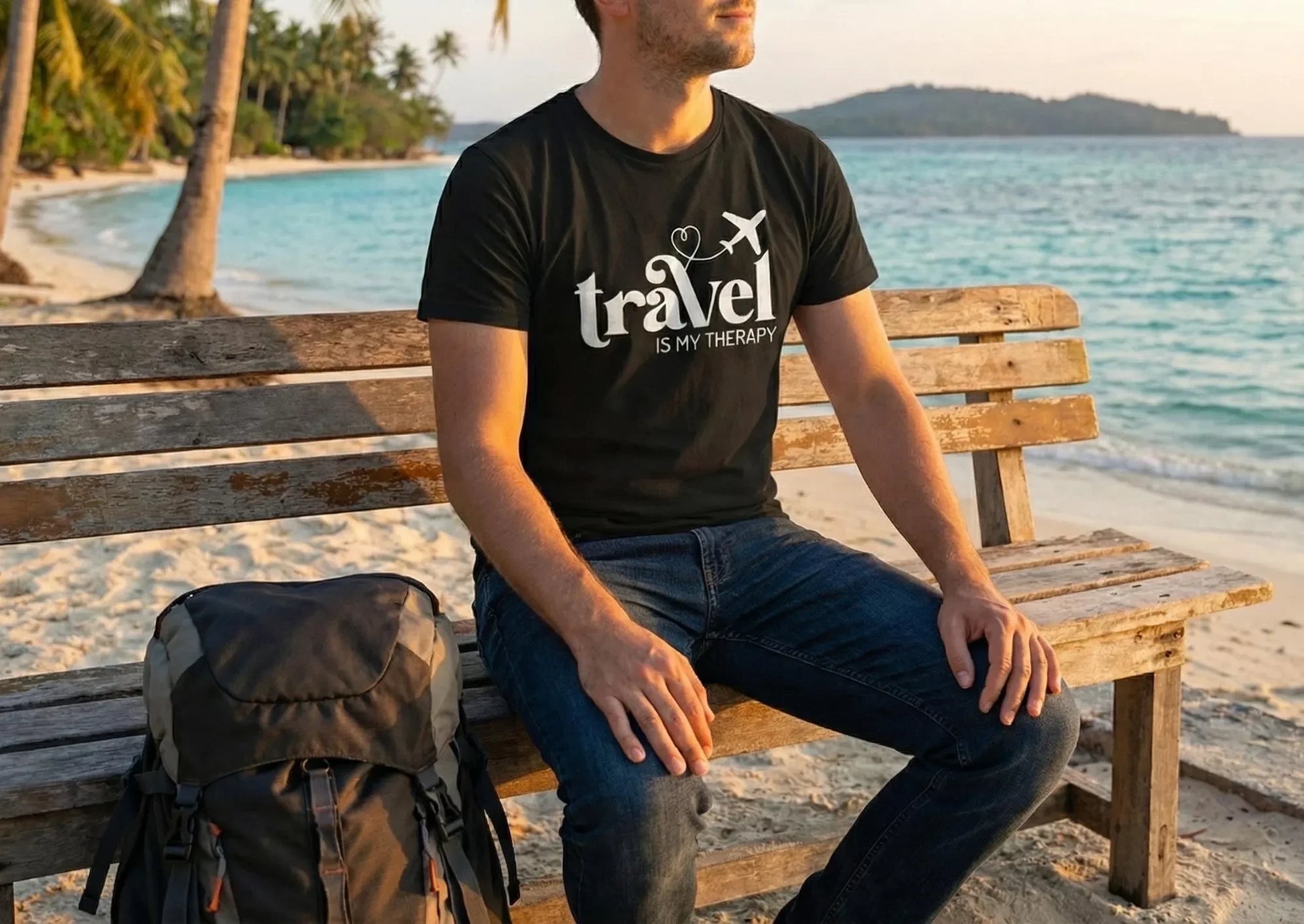 Man sitting on a bench by the beach wearing a black t-shirt with 'travel is my therapy' text.